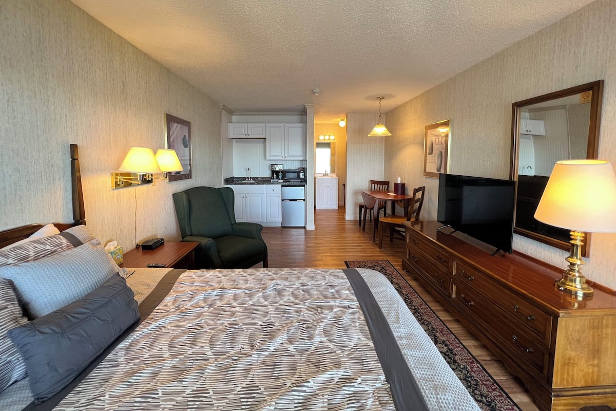 Une chambre d’hôtel avec un lit, un bureau, une chaise, une télévision, une commode et un coin cuisine à l’arrière-plan.