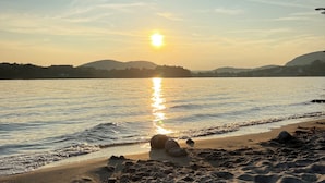 Une plage avec un coucher de soleil sur l’eau, un rivage sablonneux et des collines lointaines.
