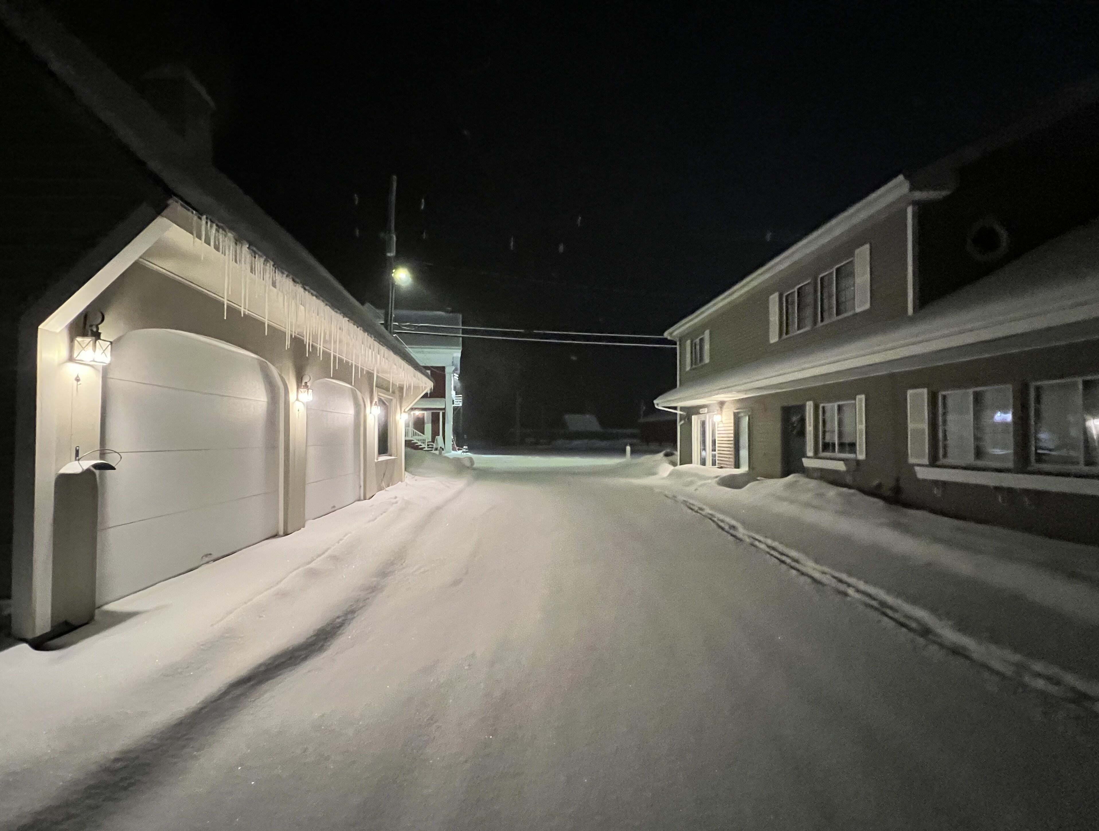 A snow-covered street at night with houses on either side, illuminated by streetlights and building lights.