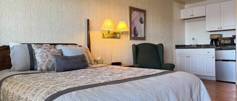 A hotel room with a large bed, a green armchair, a kitchenette, and a wall-mounted lamp.