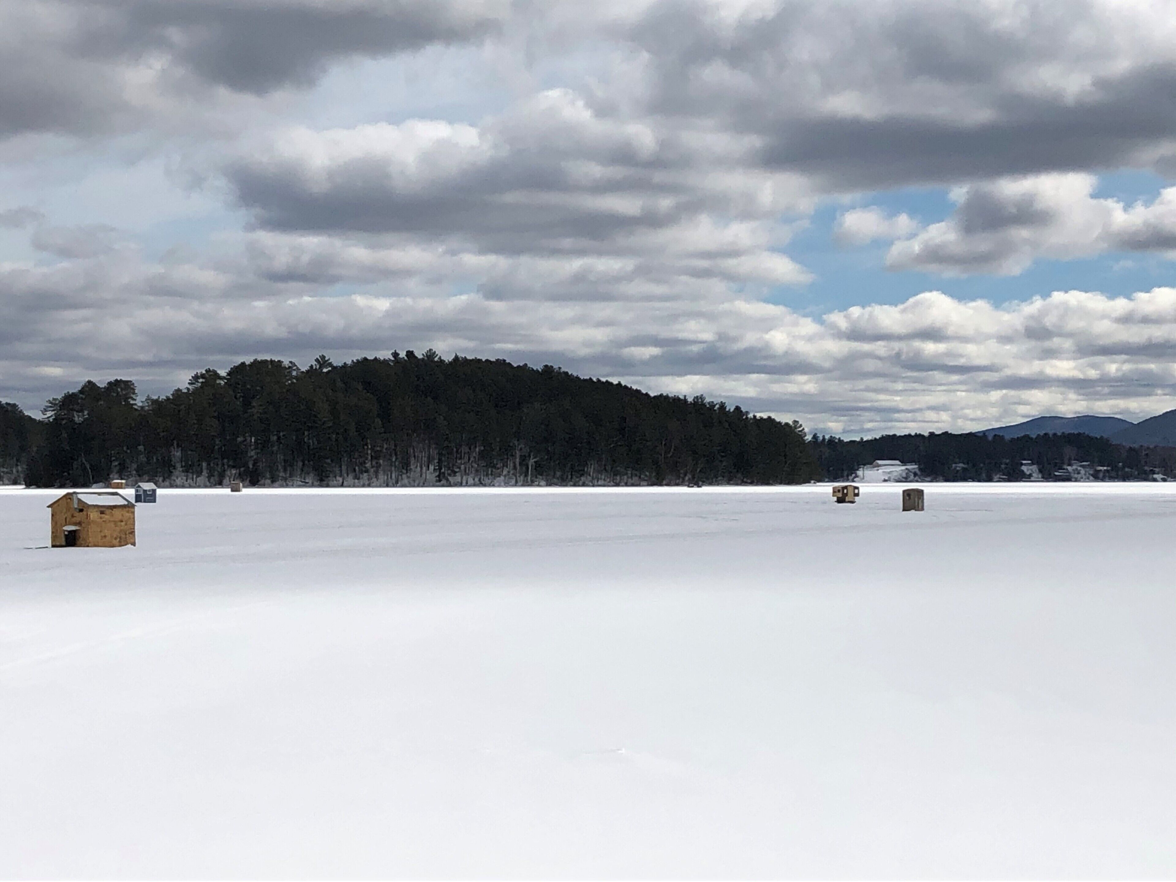 A snowy landscape with a small wooden cabin and a hay bale in the foreground, a forested hill in the background, and a partly cloudy sky.