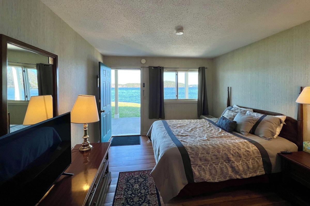 Une chambre d’hôtel avec un lit, des lampes de chevet, un bureau avec une lampe, un miroir et une vue sur la mer par la fenêtre.