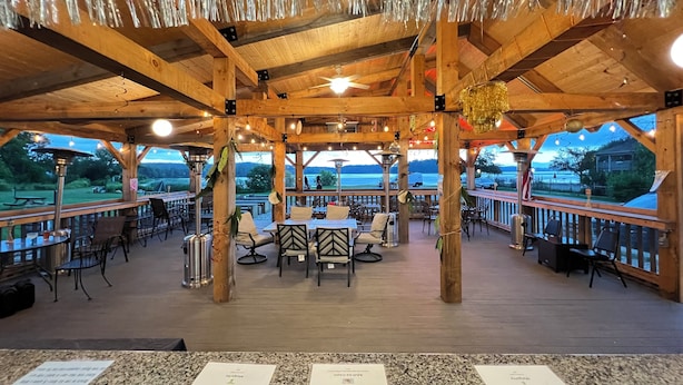 Une terrasse extérieure en bois, dotée de tables et de chaises, entourée d’une rambarde et offrant une vue sur une étendue d’eau.