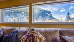 Ein Wohnzimmer mit einer Couch und zwei Fenstern mit Blick auf eine Berglandschaft.
