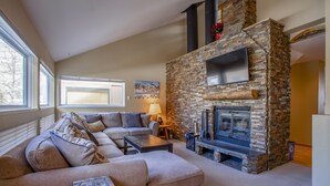 Ein gemütliches Wohnzimmer mit Kamin aus Stein, beigen Sofas, einem Couchtisch und einem an der Wand montierten Fernseher.