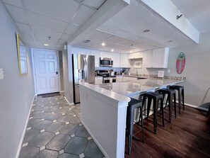 A modern kitchen with a central island, stainless steel appliances, and a breakfast bar with stools. - Slice of Salty Life!  Beachfront Condo, Spectacular Balcony, Beach Service, Pool (Panama City Beach)