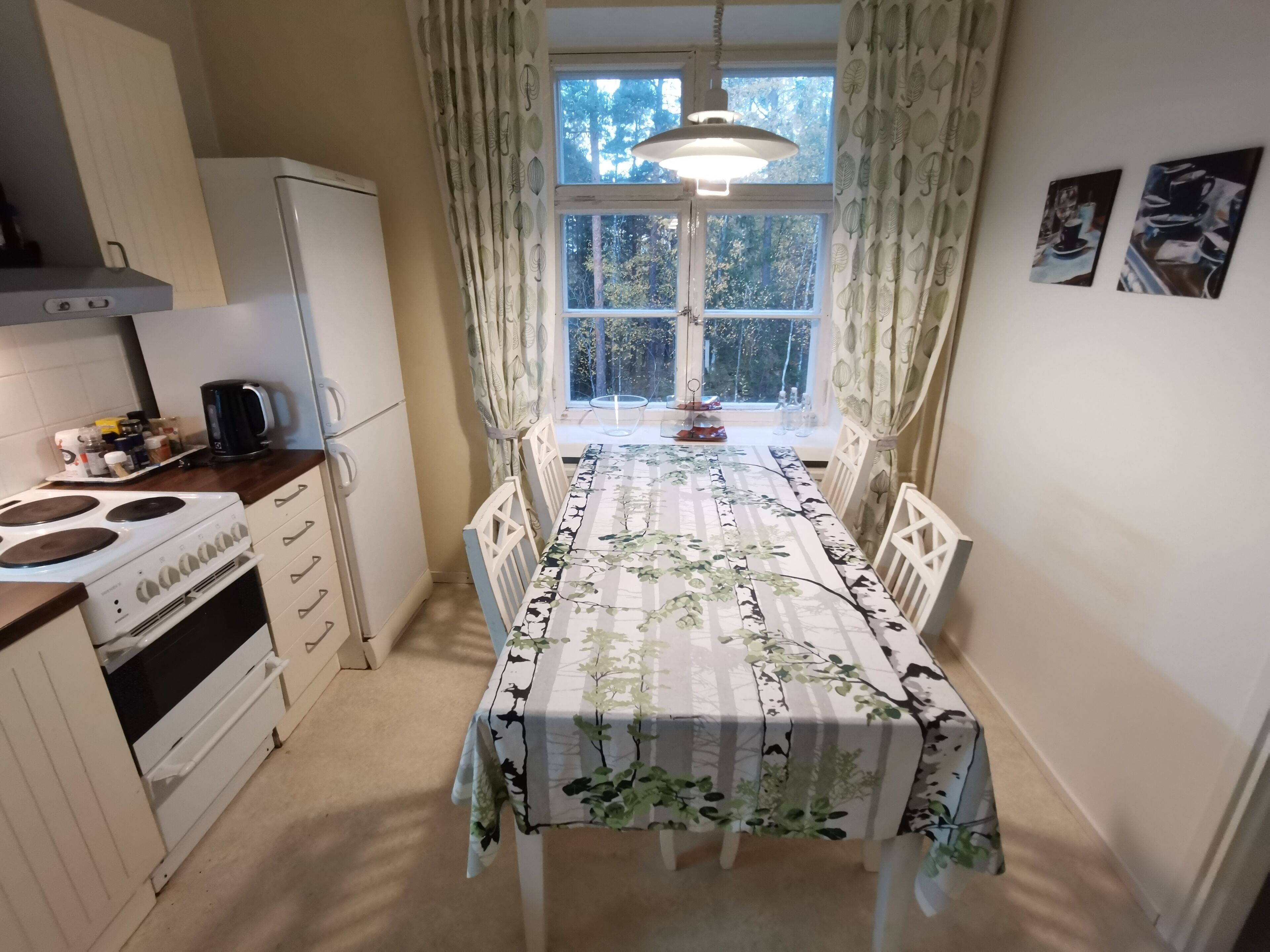 Un coin repas avec une table recouverte d’une nappe à motifs floraux, des chaises blanches, un poêle blanc et un réfrigérateur.
