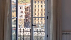 Vista desde una ventana con baranda metálica, que muestra un paisaje urbano con edificios y una colina.