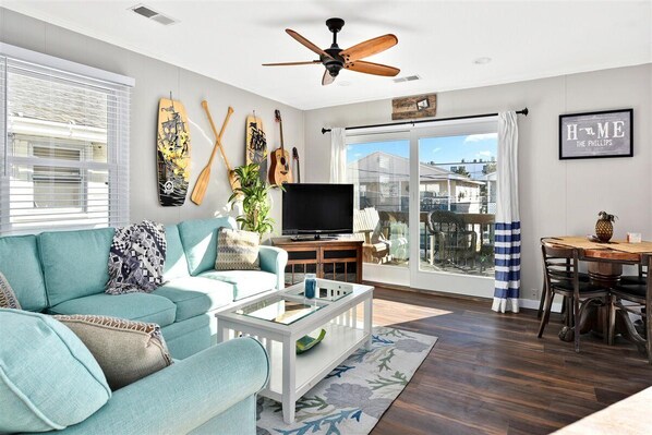 A modern living room with a teal sofa, a glass coffee table, and a TV on a wooden stand. There are guitars on the wall and a view of the outside through large windows.
