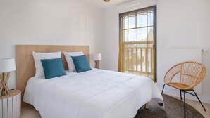 A bedroom with a large bed, two bedside tables with lamps, a wicker chair, and a window with a curtain.