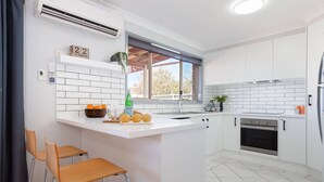 A modern kitchen with white cabinetry, a central island, a built-in oven, and a window providing natural light. - Blue Bay - Close to Fingal Bay Beach. Mid Week Stay Special Now On. (Fingal Bay)