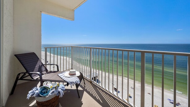 Un balcon avec vue sur la plage, une chaise, une table avec un chapeau et un panier rempli de serviettes.