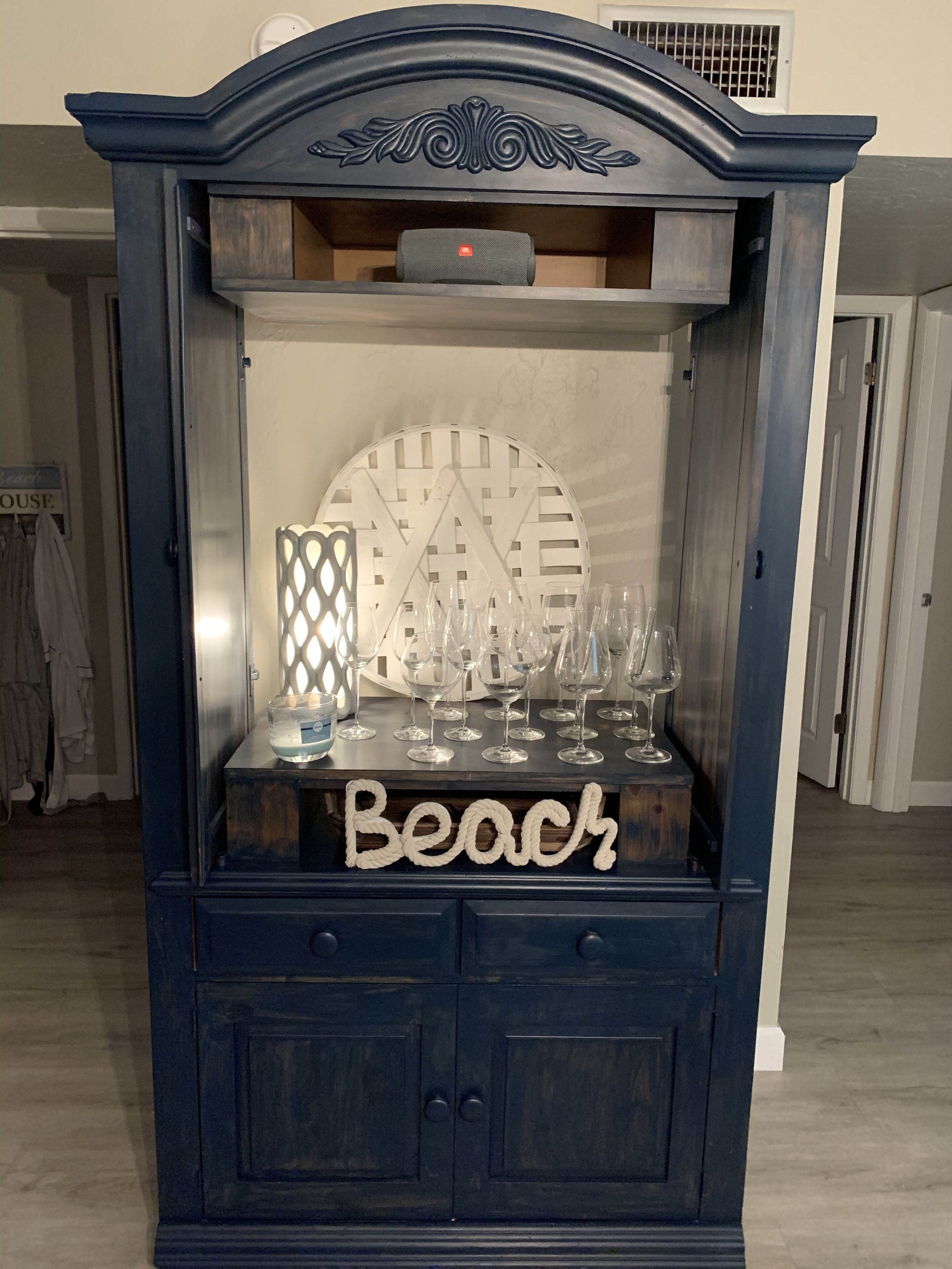 Un meuble bleu foncé, orné d’un élément décoratif sur le dessus, sur lequel est apposée une pancarte indiquant « Plage », ainsi qu’une collection de verres à vin et un chandelier.