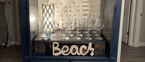 Un meuble bleu foncé, orné d’un élément décoratif sur le dessus, sur lequel est apposée une pancarte indiquant « Plage », ainsi qu’une collection de verres à vin et un chandelier.