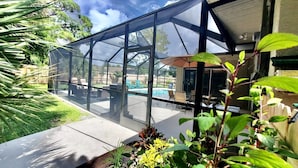 Un patio con paredes de vidrio, piscina, muebles de exterior y abundante vegetación.