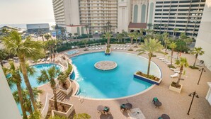 A resort pool area with multiple lounge chairs and a central circular pool, surrounded by palm trees and a high-rise building in the background. - 'Ocean Oasis' PCB Condo w/ Balcony & Beach Views! (Panama City Beach)