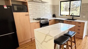 Une cuisine moderne avec un îlot en marbre, des armoires en bois, un réfrigérateur noir, une cuisinière et une fenêtre donnant sur les arbres.
