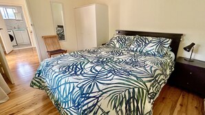 Une chambre à coucher avec un lit, un plancher en bois, une table de chevet, une lampe, une chaise, une armoire et une salle de bain visible au fond.