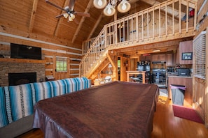 Interior de una cabaña de madera con mesa de comedor, cocina, chimenea y un balcón con baranda de madera.