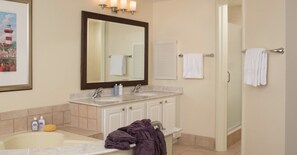 A bathroom with a large mirror, two sinks, a bathtub, and a shower. There are towels hanging on the wall and a framed picture above the mirror.