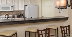 A kitchen with white cabinets, a microwave, a refrigerator, a coffee maker, and a kettle on the counter. There are three bar stools and a glass table.