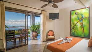 A hotel room with a bed, a television, a painting of a parrot, a chair, and a view of the ocean. - Villa Costa Mar Ocean View Guard Gated Community (Playa Hermosa, Carrillo)