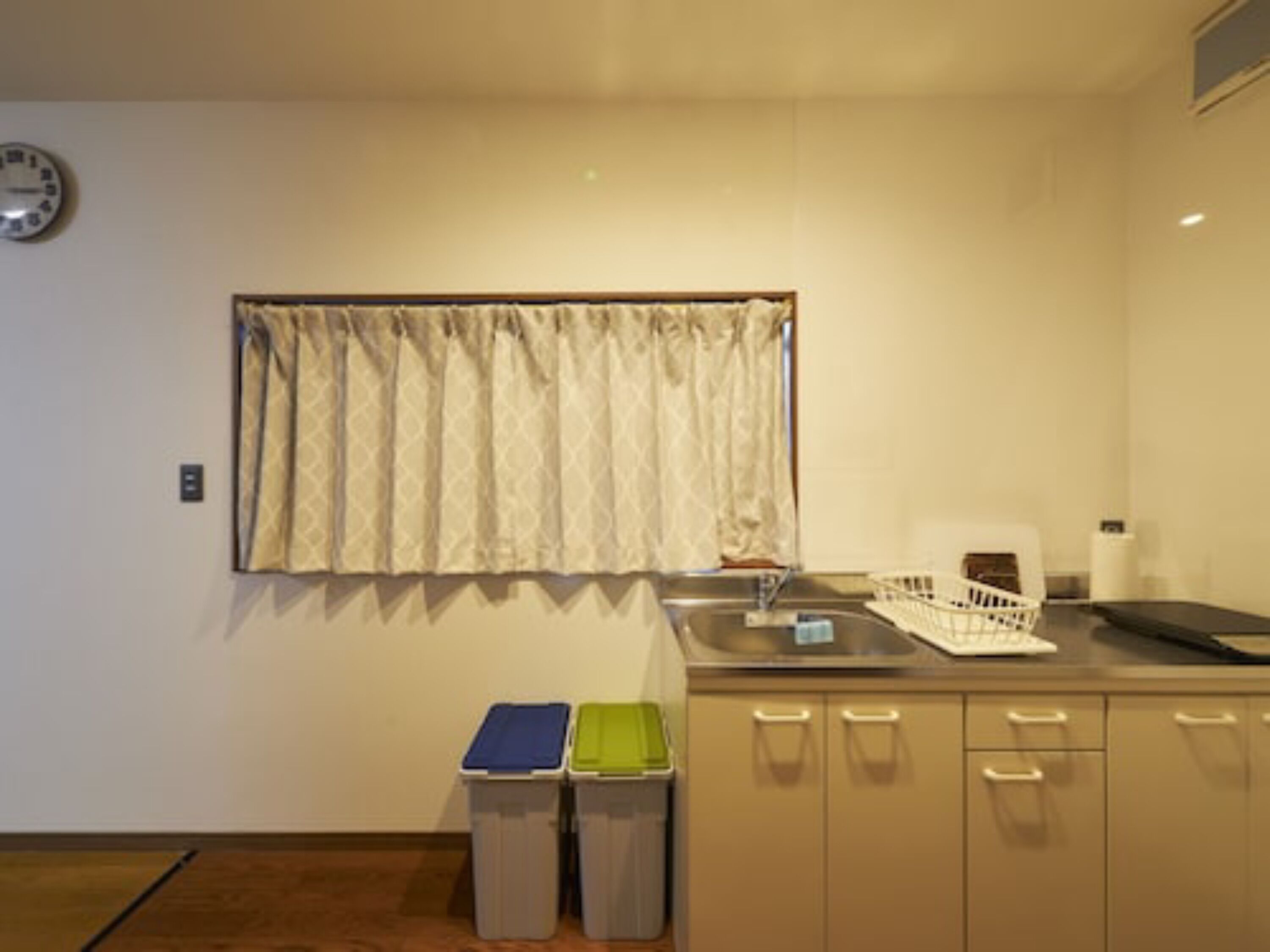 A small kitchenette with a window, a clock, and two trash bins.