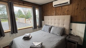 A bedroom with a large bed, a nightstand, an air conditioning unit, and a view through the window.