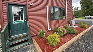 A red building with a green door labeled "Apartment 1," a small garden with mulch and plants, and a white car parked in the background.