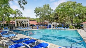 A swimming pool with blue lounge chairs, a building with a red roof, and trees in the background.