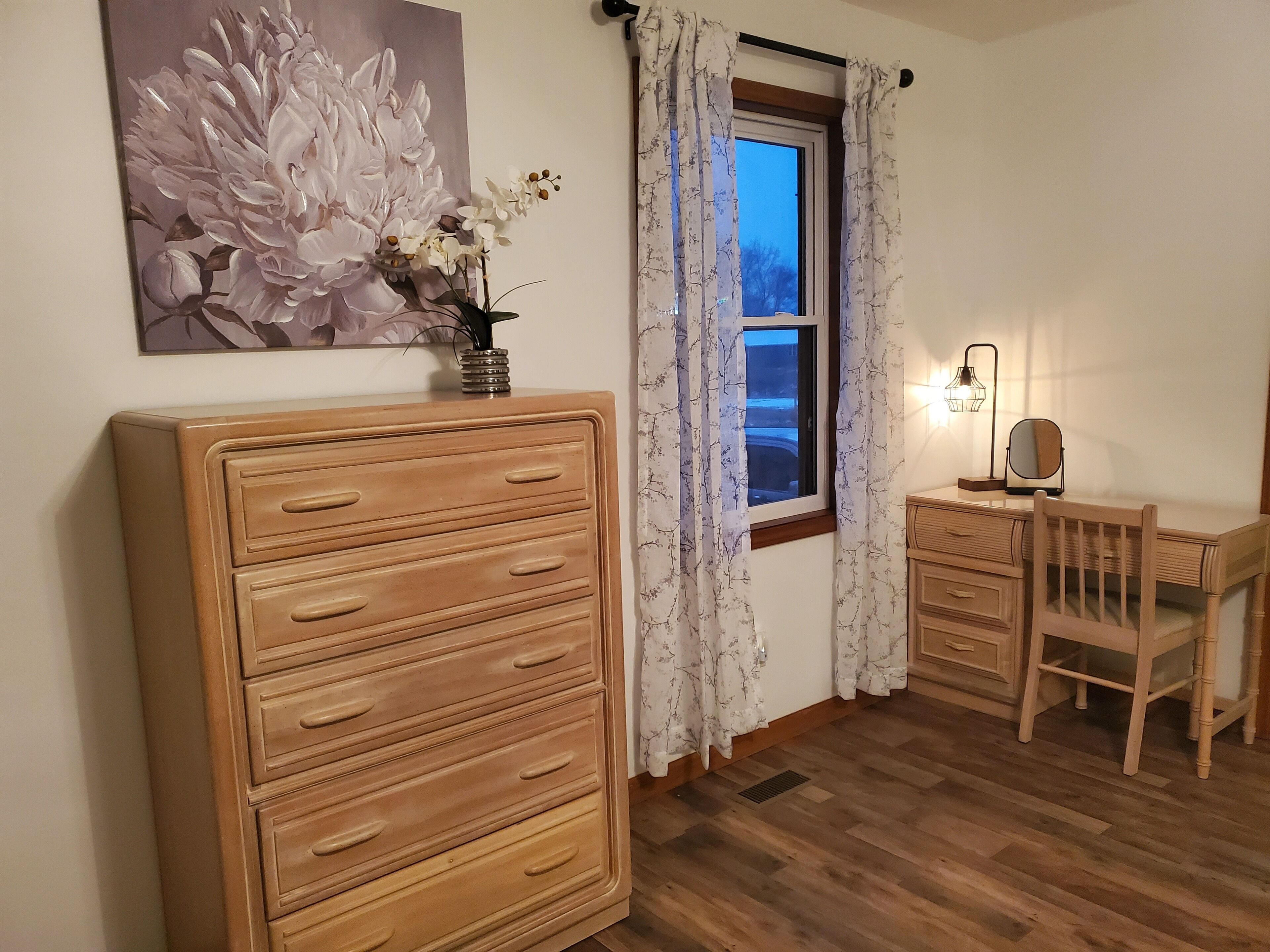Una habitación con un amplio aparador de madera, un escritorio con silla, una ventana con cortinas, una lámpara fijada a la pared y un cuadro con motivos florales.