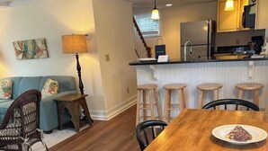 Ein Essbereich mit einem Holztisch und Stühlen, im Hintergrund eine Küche und ein Wohnzimmer mit einer Couch und einer Lampe.