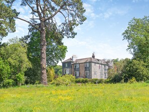 A large stone building with multiple windows and a flag, surrounded by lush greenery and a field of yellow flowers.