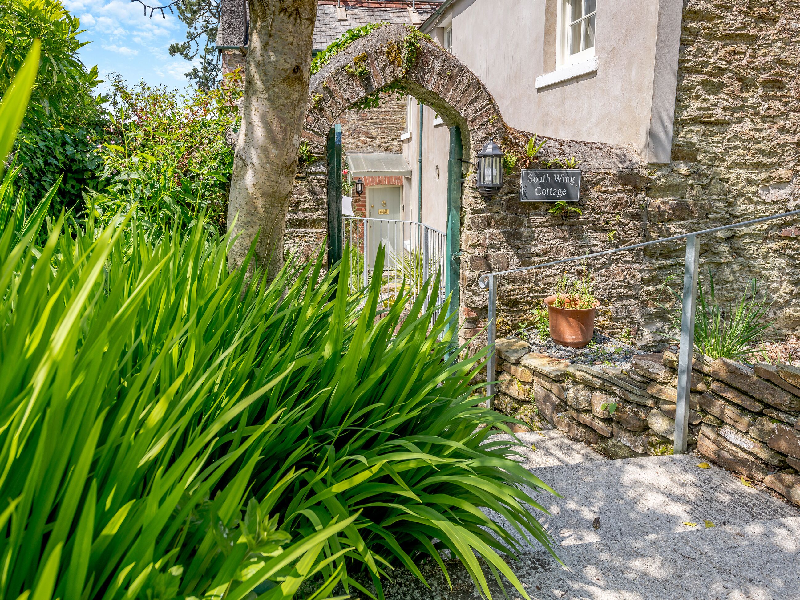 A stone cottage with a sign reading 'South Wing Cottage,' surrounded by lush greenery and a metal gate.