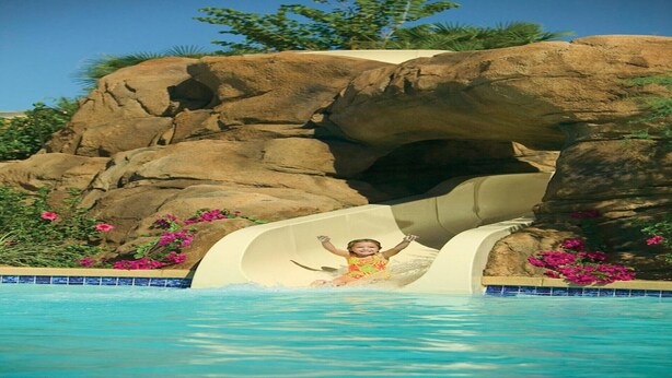 Un niño deslizándose por un tobogán acuático hacia una piscina, con un fondo rocoso y flores rosas.