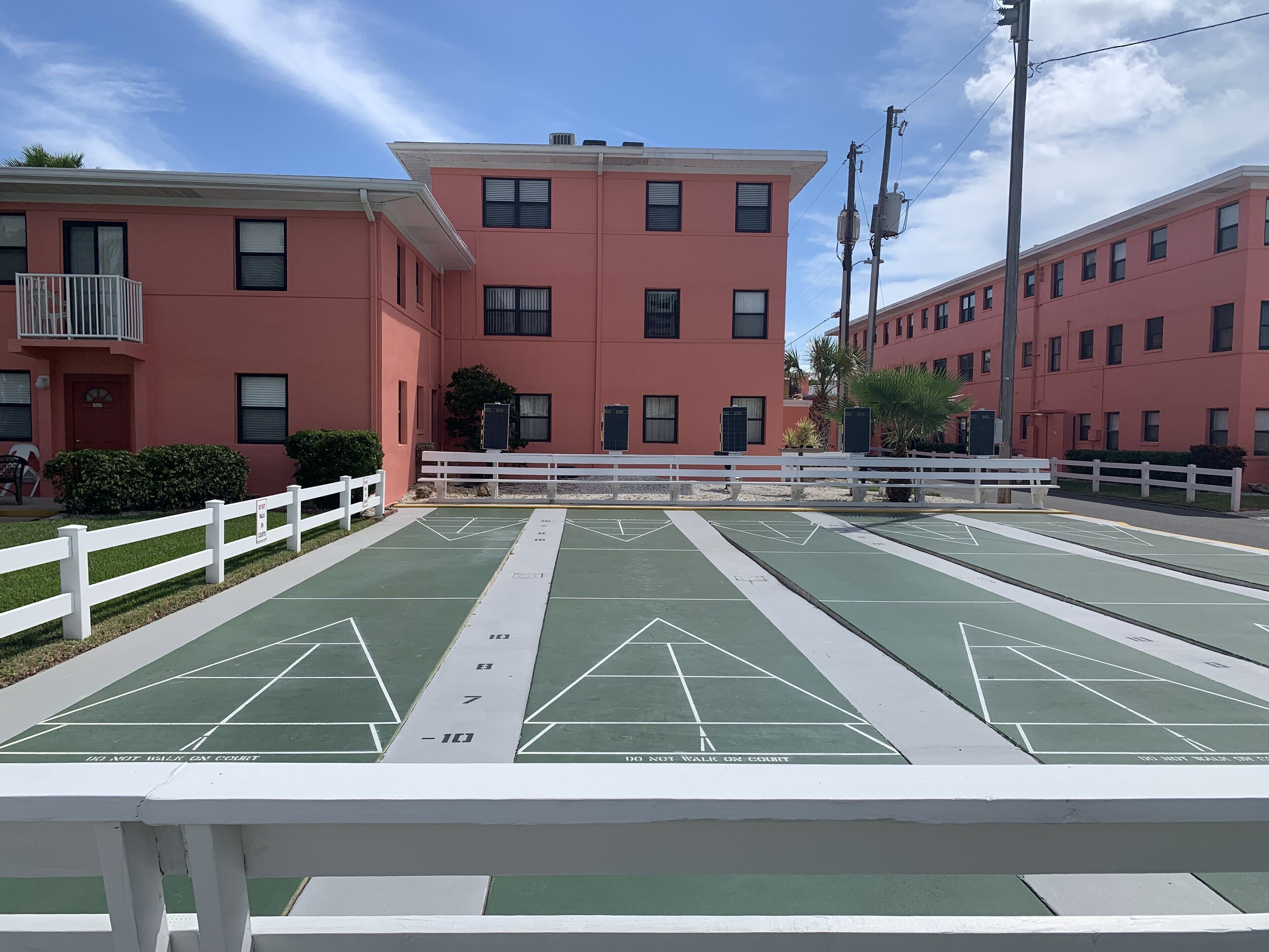 Una cancha de shuffleboard con una valla blanca en primer plano, un edificio rosa con varias ventanas y un balcón al fondo, y un cielo azul despejado.