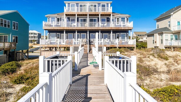 A multi-story beachfront house with a wooden deck leading to the entrance, surrounded by sandy dunes and other residential buildings. - Oceanfront stunner in Emerald Isle (Emerald Isle)