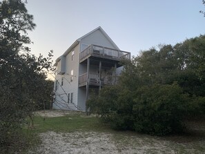 A multi-story building with a balcony on the second floor, surrounded by dense foliage. - A private, wooded home in Emerald Isle, NC, 5th row from the beach (5 min walk) (Emerald Isle)