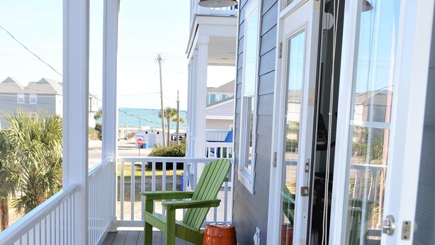 Eine überdachte Veranda mit einem grünen Adirondack-Sessel und einem kleinen Tisch, einer Schiebetür und Blick auf den Strand sowie weitere Gebäude.