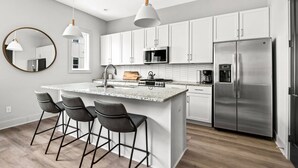 A modern kitchen with white cabinets, a stainless steel refrigerator, a built-in oven, and a granite countertop. There are three bar stools and pendant lights above the island. - H18 ~ Large Condo w/ Private Rooftop with Downtown Views ~ Garage Parking ~ 9 Minute to Broadway (Nashville)