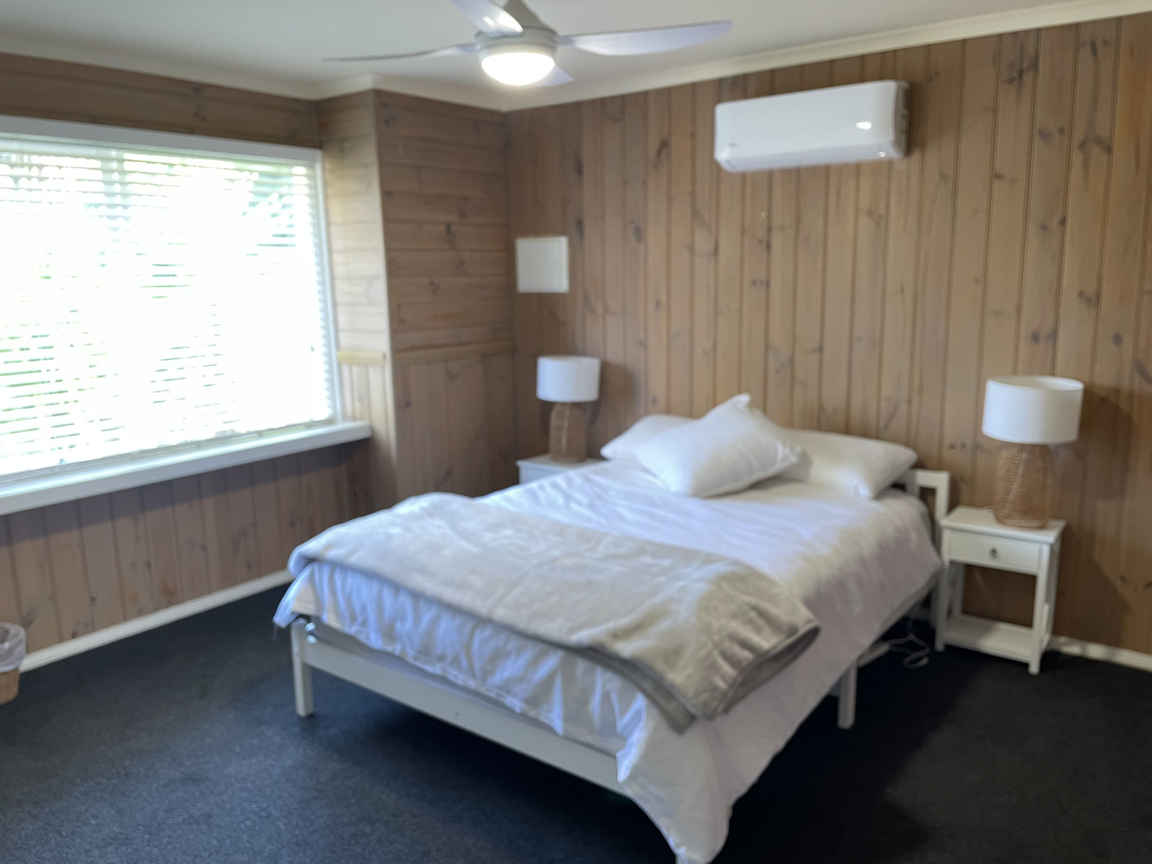 Une chambre à coucher comprenant un lit, deux tables de chevet, un ventilateur de plafond et une fenêtre avec des stores.