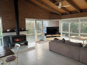 A cozy living room with a fireplace, a television, a sofa, and large windows. - Spacious ocean view beach house - sleeps 12 (Smiths Beach)