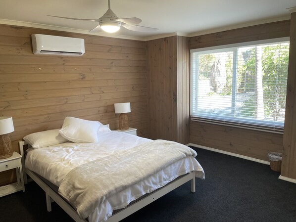 A bedroom with a bed, two bedside tables with lamps, a ceiling fan, an air conditioning unit, and a window with blinds. - Spacious ocean view beach house - sleeps 12 (Smiths Beach)