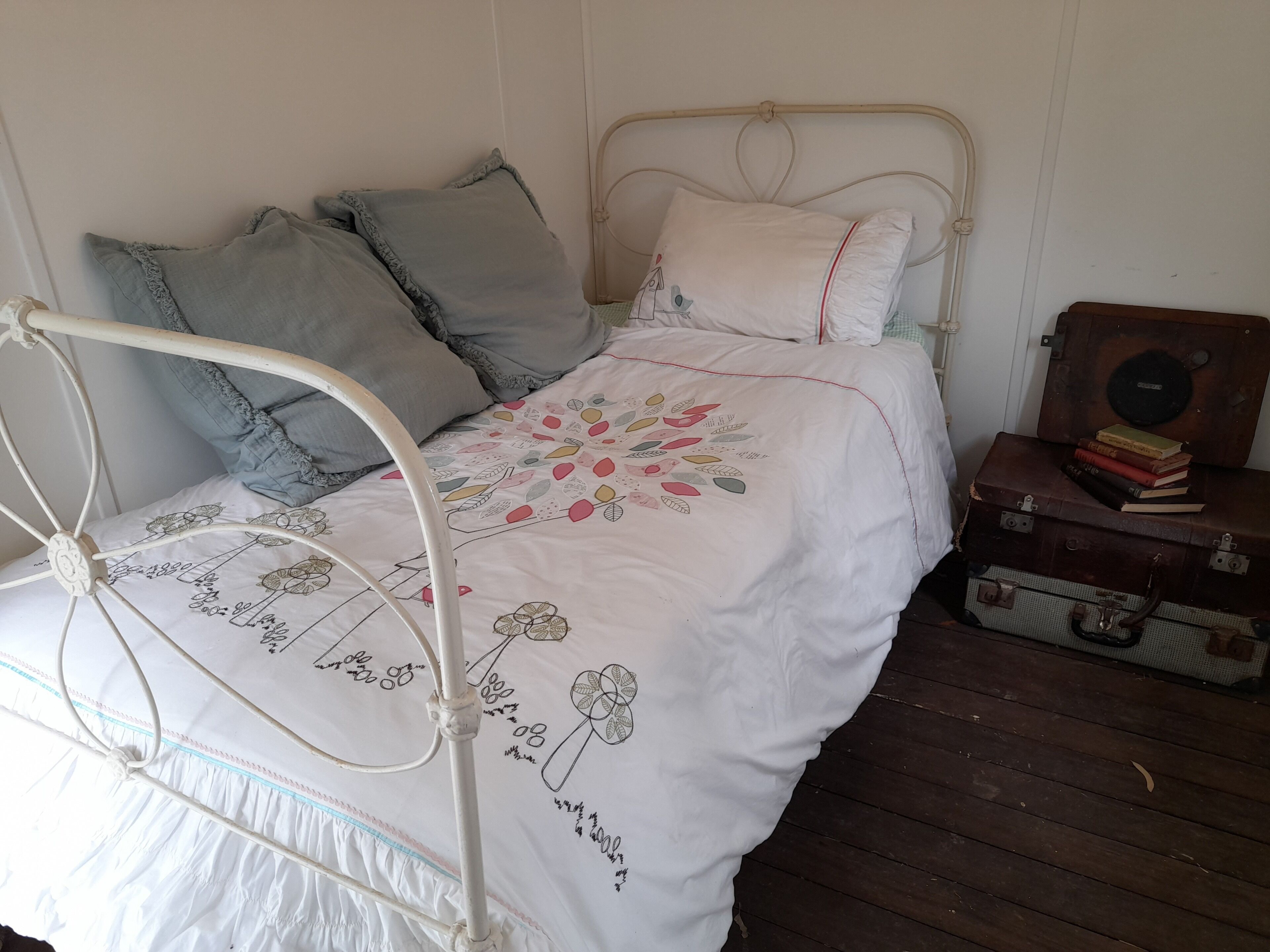 Una cama con estructura metálica, ropa de cama blanca con bordados florales y una mesita de noche de madera con libros.