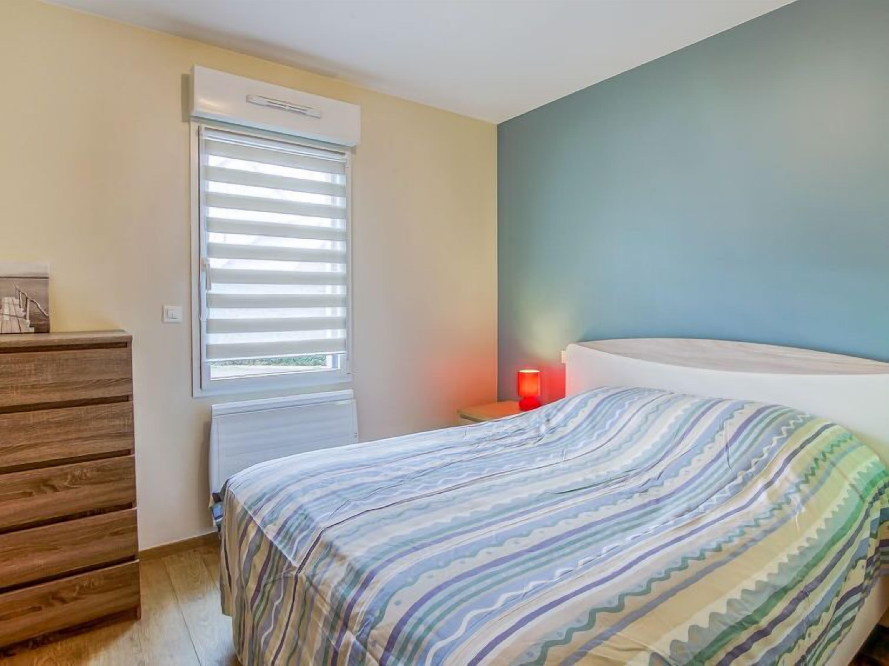 A bedroom with a bed, a dresser, a window with blinds, and a nightstand with a lamp.