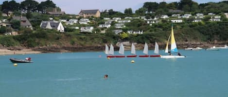A coastal area with sailboats on the water, a beach, and houses in the background.