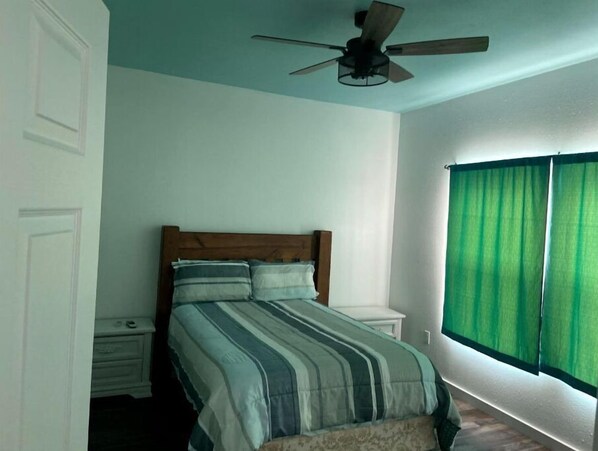 Een slaapkamer met een bed, een plafondventilator, een nachtkastje en een raam met groene gordijnen.
