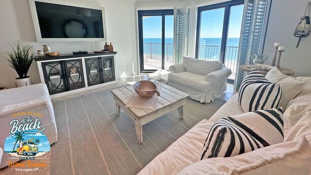 A spacious living room with a large TV, white furniture, a view of the ocean, and a balcony with shutters. - Extra Large Balcony Phoenix VII 3/2 Flash Sale (Orange Beach)