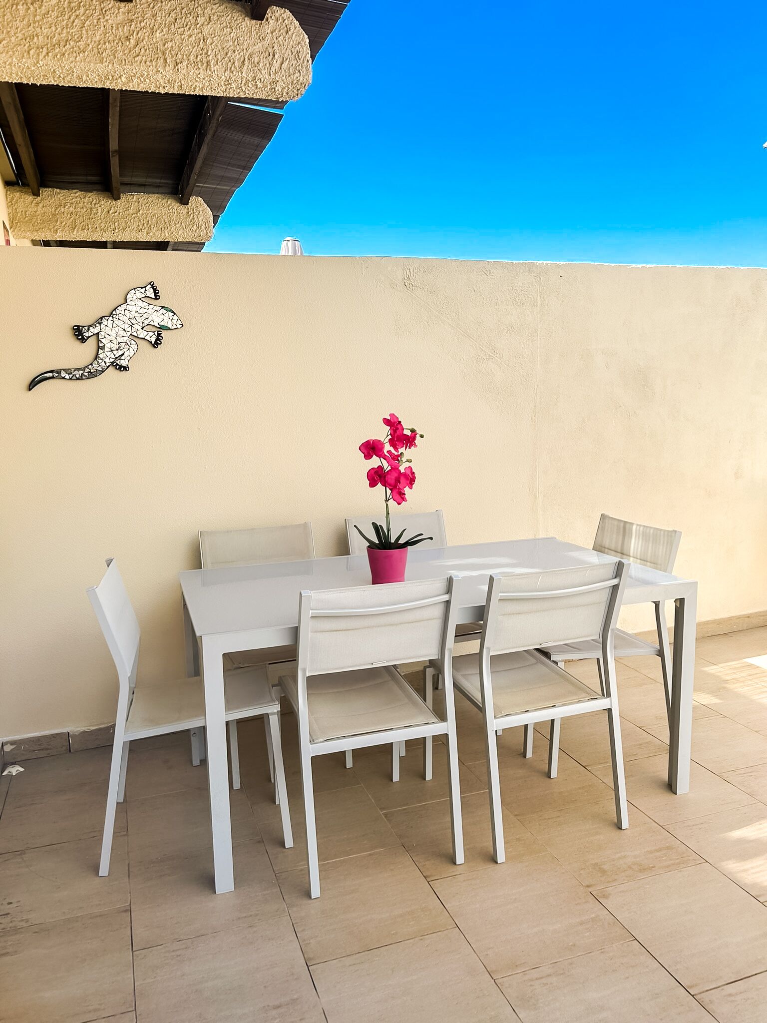 A white outdoor dining set with chairs and a table, a potted plant, and a decorative wall piece against a plain wall.