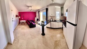 A modern kitchen with white cabinets, a black countertop, and a built-in oven. There is a dining area with a table and chairs, and a pink wall in the background. - Beautiful three bedroom apartment with shared swimming pool and WIFI (Puerto de Mogan)